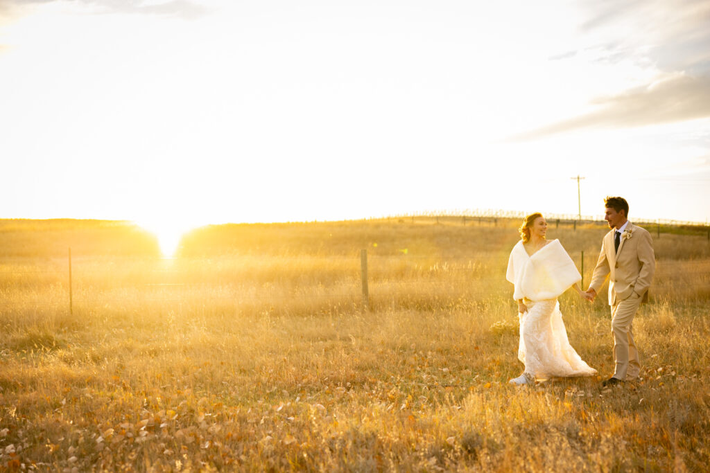 Newlywed couple walking in a prairie at sunset, with the golden sky in the background.