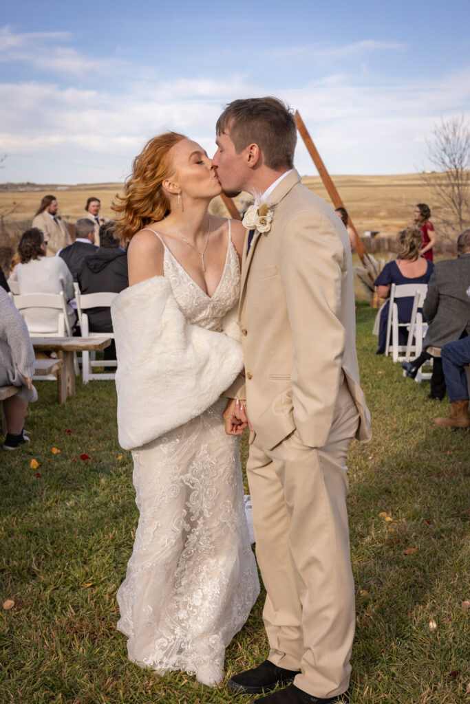 Couple kissing amidst a countryside wedding ceremony with guests seated in the background.
